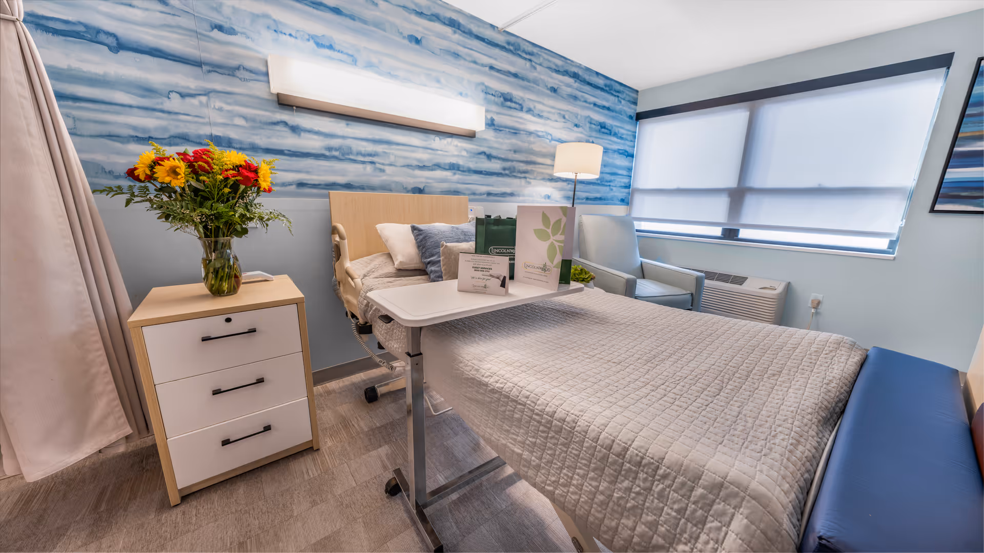 A bright and clean patient room in a rehabilitation and healthcare center featuring a hospital bed with a white quilted blanket, a bedside table with a vase of colorful flowers, a chair near the window with blinds partially closed, and a blue and white abstract wall design behind the bed.