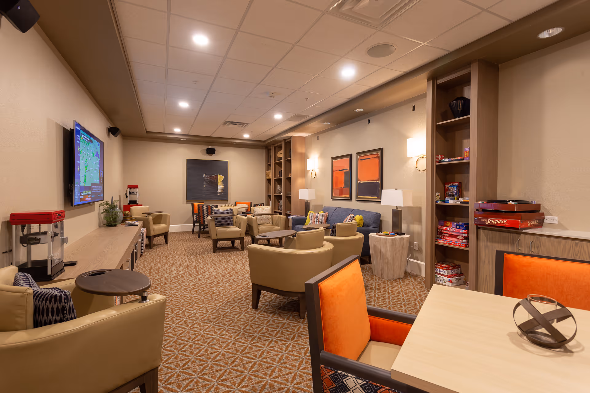 A cozy senior living common area with beige walls and carpeted floor. The room features multiple comfortable armchairs and sofas arranged around small tables. There is a flat-screen TV mounted on the left wall, a popcorn machine on a long wooden cabinet, and shelves filled with board games like Scrabble. The lighting is warm with ceiling lights and wall sconces, and colorful abstract paintings decorate the walls.
