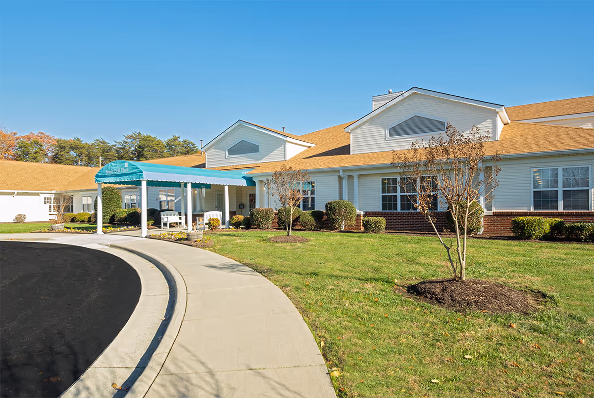 Exterior view of Cardinal Senior Living at Danville showing a single-story building with white siding and a tan roof. There is a covered entrance with a teal awning, a curved driveway, and a well-maintained lawn with small trees and shrubs under a clear blue sky.
