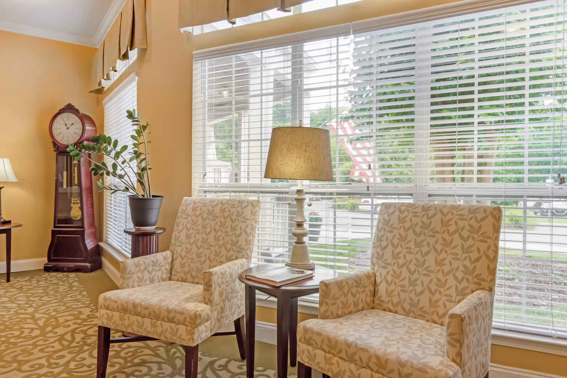 Two patterned armchairs flank a small table with a lamp in a sunlit sitting area by large windows and a grandfather clock.