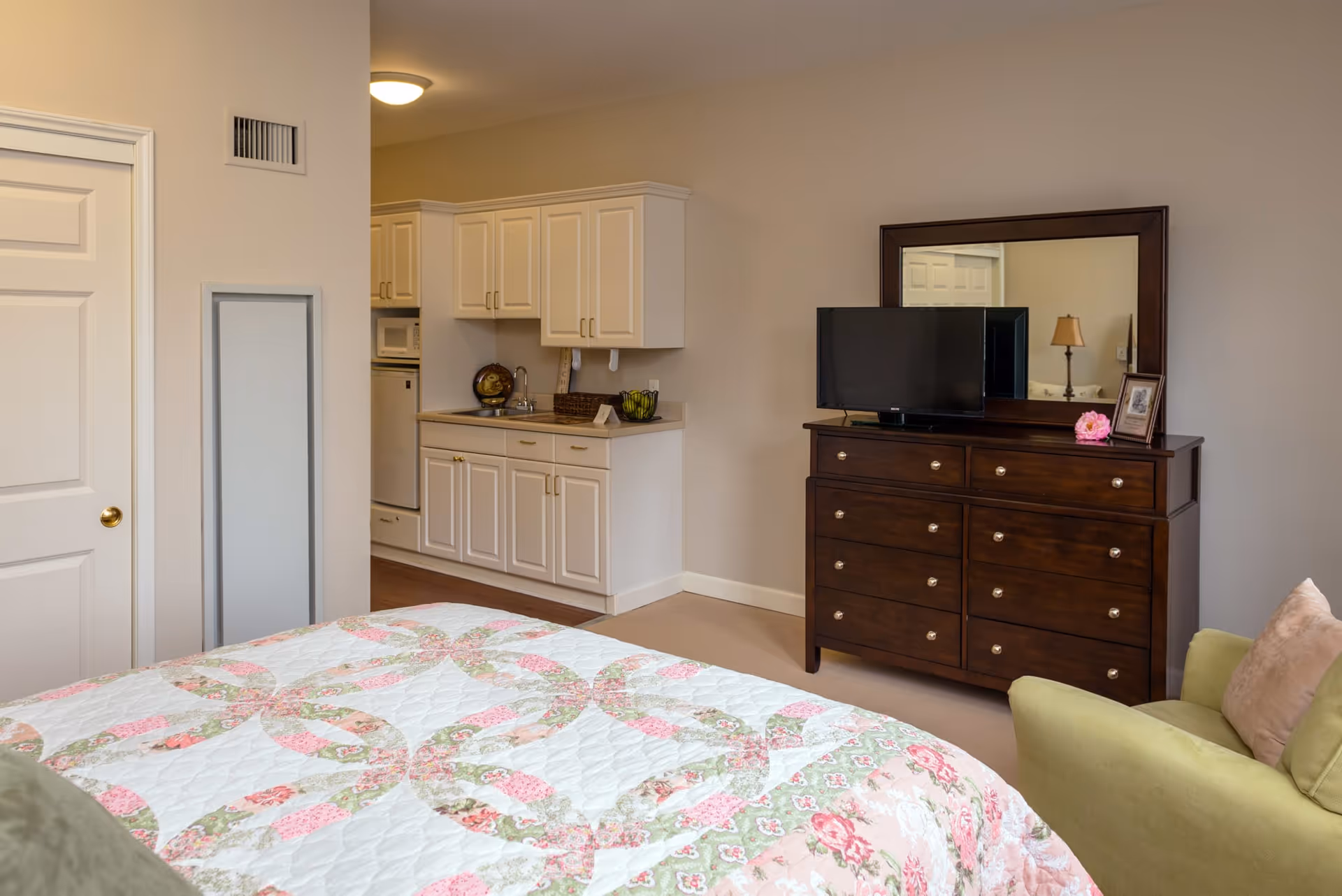 A cozy bedroom area with a floral patterned quilt on the bed, a green armchair with a beige pillow, a dark wooden dresser with a mirror and a TV on top, and a small kitchenette with white cabinets, a sink, a microwave, and a mini fridge.