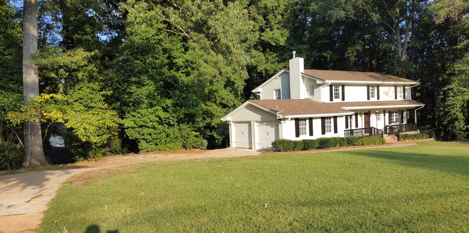 A two-story white house with a brown roof and black shutters surrounded by green trees and a well-maintained lawn. The house has a double garage and a front porch with a ramp and stairs.