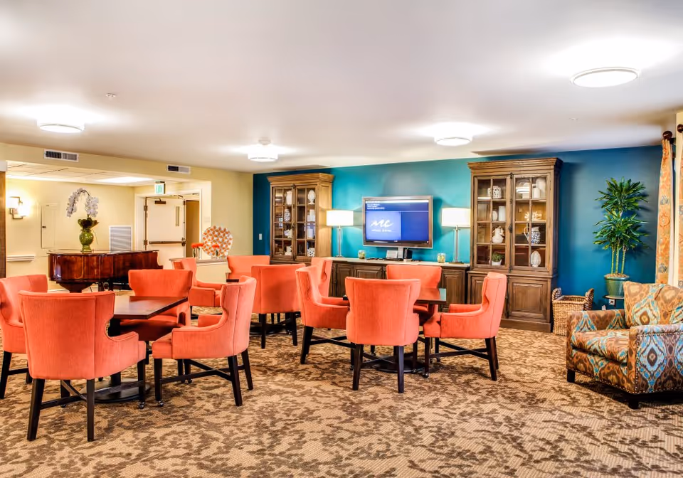 A bright and spacious common area with coral upholstered chairs arranged around dark wooden tables on a patterned carpet. The room features a teal accent wall with two wooden display cabinets and a flat-screen TV mounted between them. There are two table lamps on a wooden console beneath the TV, a patterned armchair on the right, a large potted plant, and a grand piano in the background near the entrance.