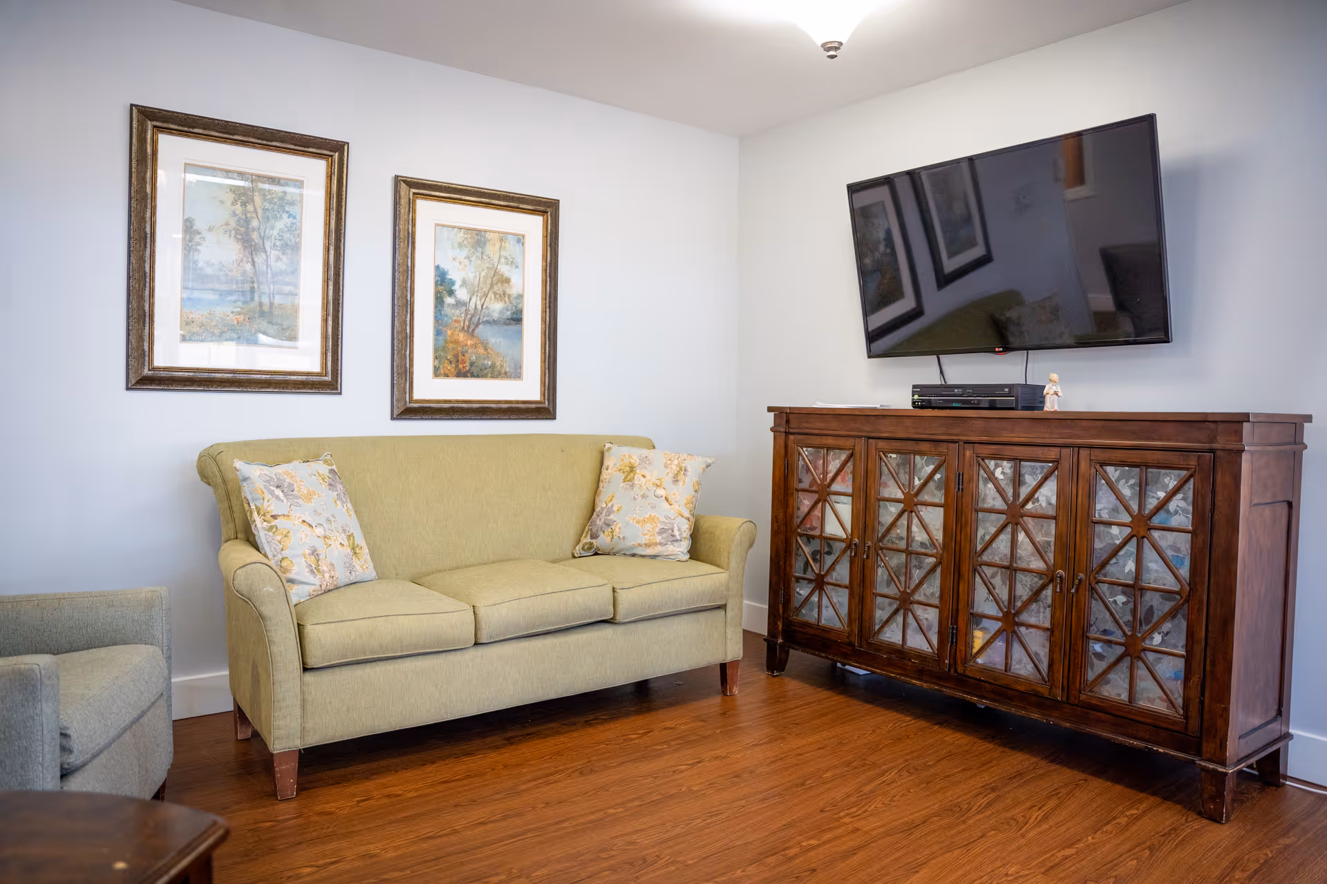 A cozy living room with a light green sofa adorned with two floral pillows, a matching armchair, two framed landscape paintings on the wall, a wooden cabinet with glass-paneled doors, and a flat-screen TV mounted above the cabinet. The room has wooden flooring and light-colored walls.