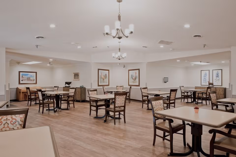 A spacious dining room with multiple tables and chairs arranged neatly on a wooden floor. The walls are decorated with framed artwork, and there are two chandeliers hanging from the ceiling providing light. The room has a clean and welcoming atmosphere.