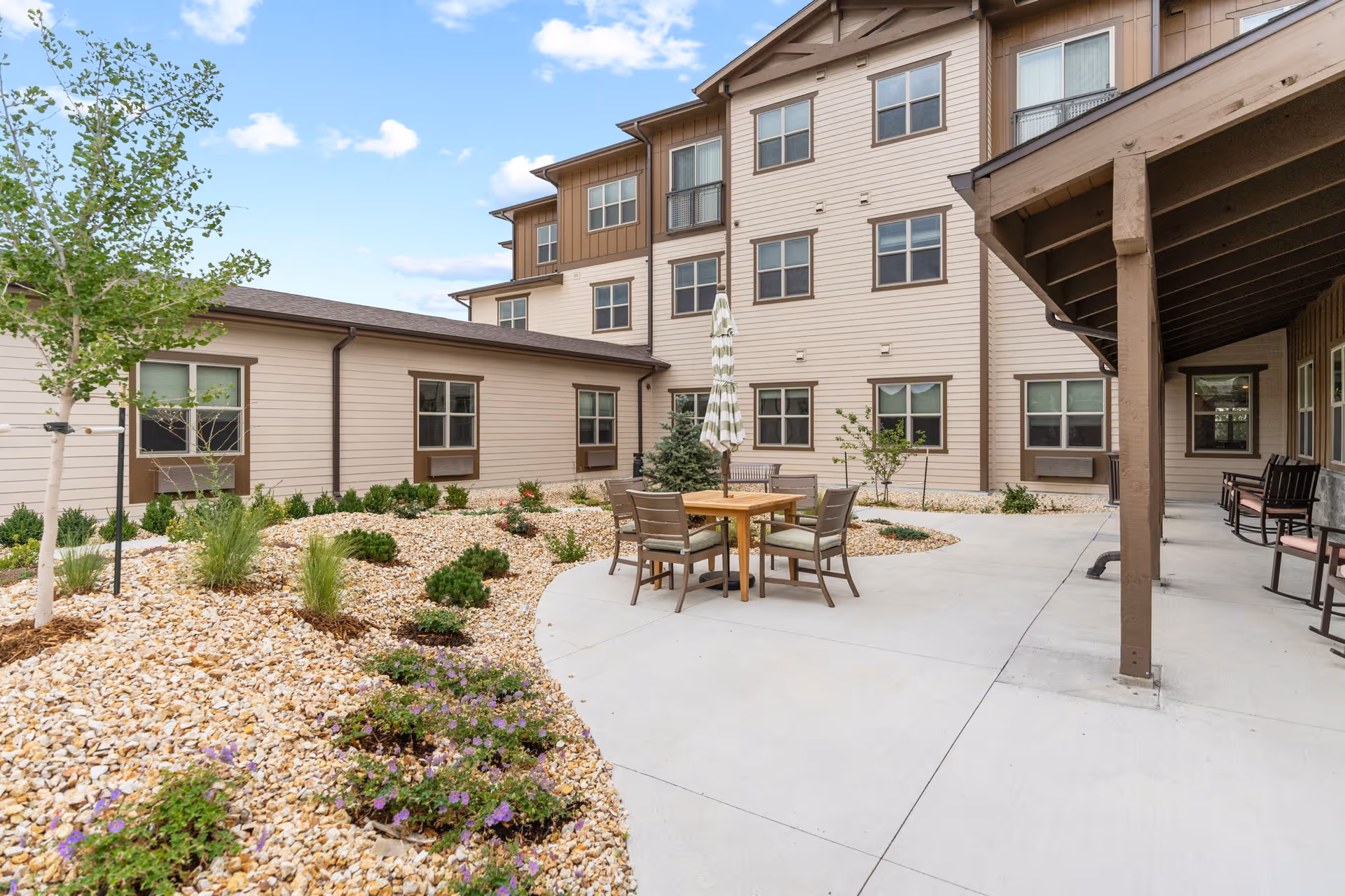 Outdoor courtyard area of a senior living facility with a small round table and four chairs under a closed umbrella. The courtyard is surrounded by a beige multi-story building with several windows. There are landscaped areas with small bushes, plants, and trees, and a concrete walkway and patio area with additional seating along the building.