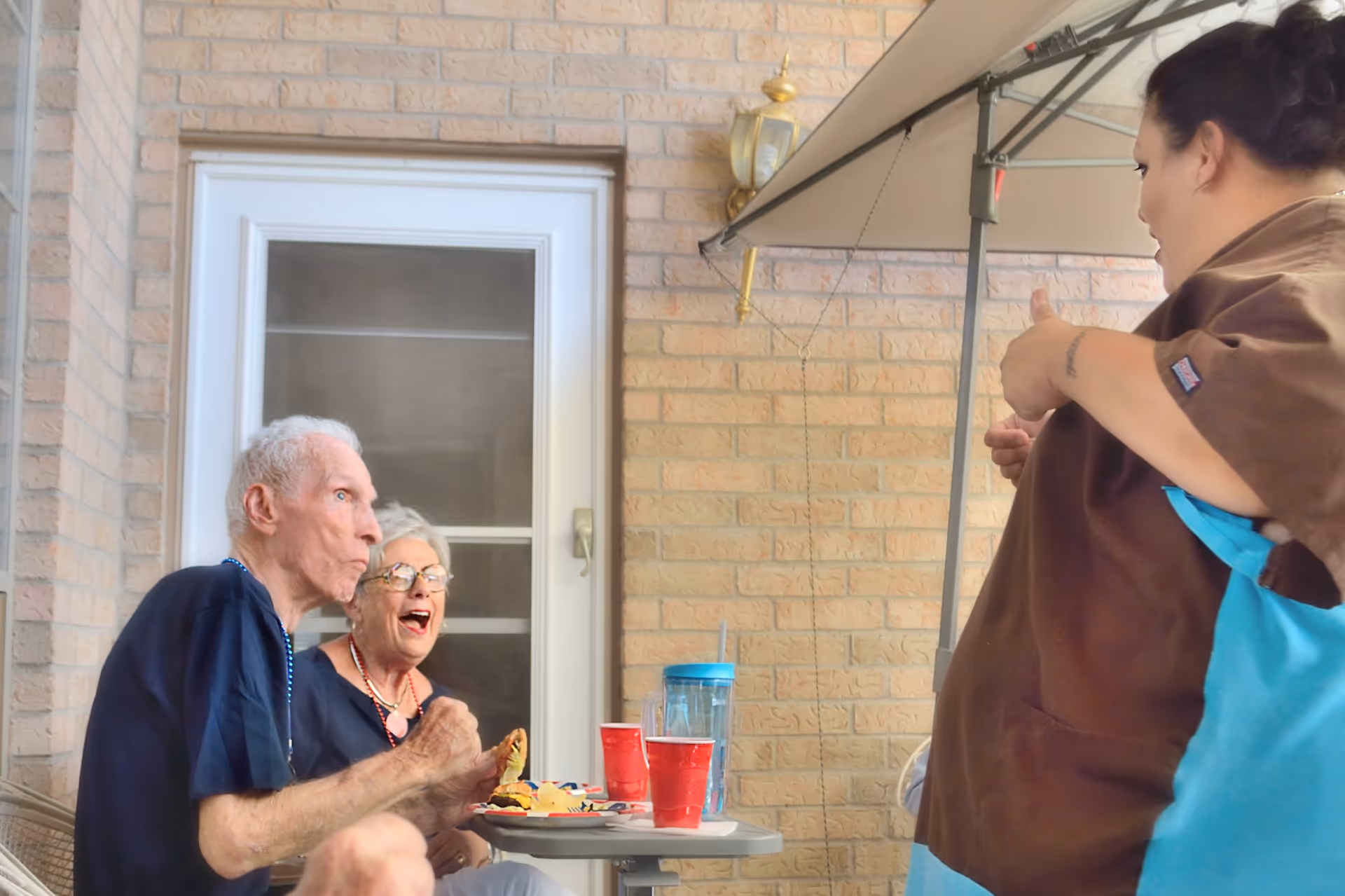 Two elderly people sitting at a small outdoor table eating snacks and interacting with a caregiver who is standing nearby giving a thumbs-up gesture. The setting is a brick patio area with a door and a canopy overhead.
