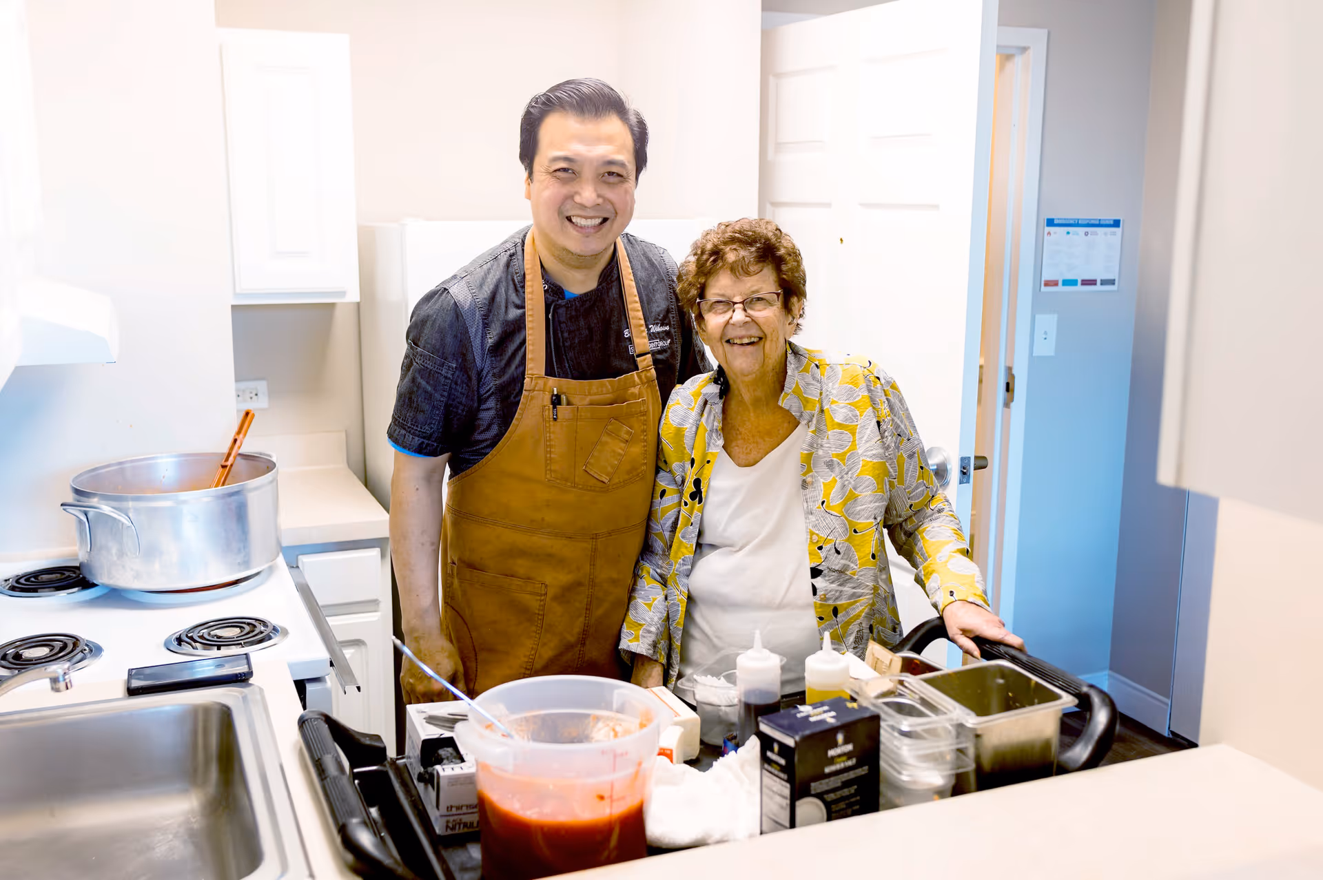 A man wearing a brown apron and a smiling elderly woman standing together in a kitchen. The man is standing near a stove with a large pot and a wooden spoon, while the woman is next to a counter with various cooking ingredients and utensils. Both are smiling and appear to be enjoying their time.