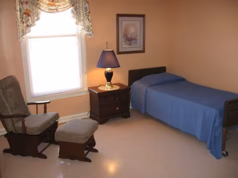 A simple bedroom with a single bed covered with a blue blanket, a wooden nightstand with a blue lamp, a framed picture on the wall, a window with floral curtains, and a cushioned rocking chair with a matching footstool.