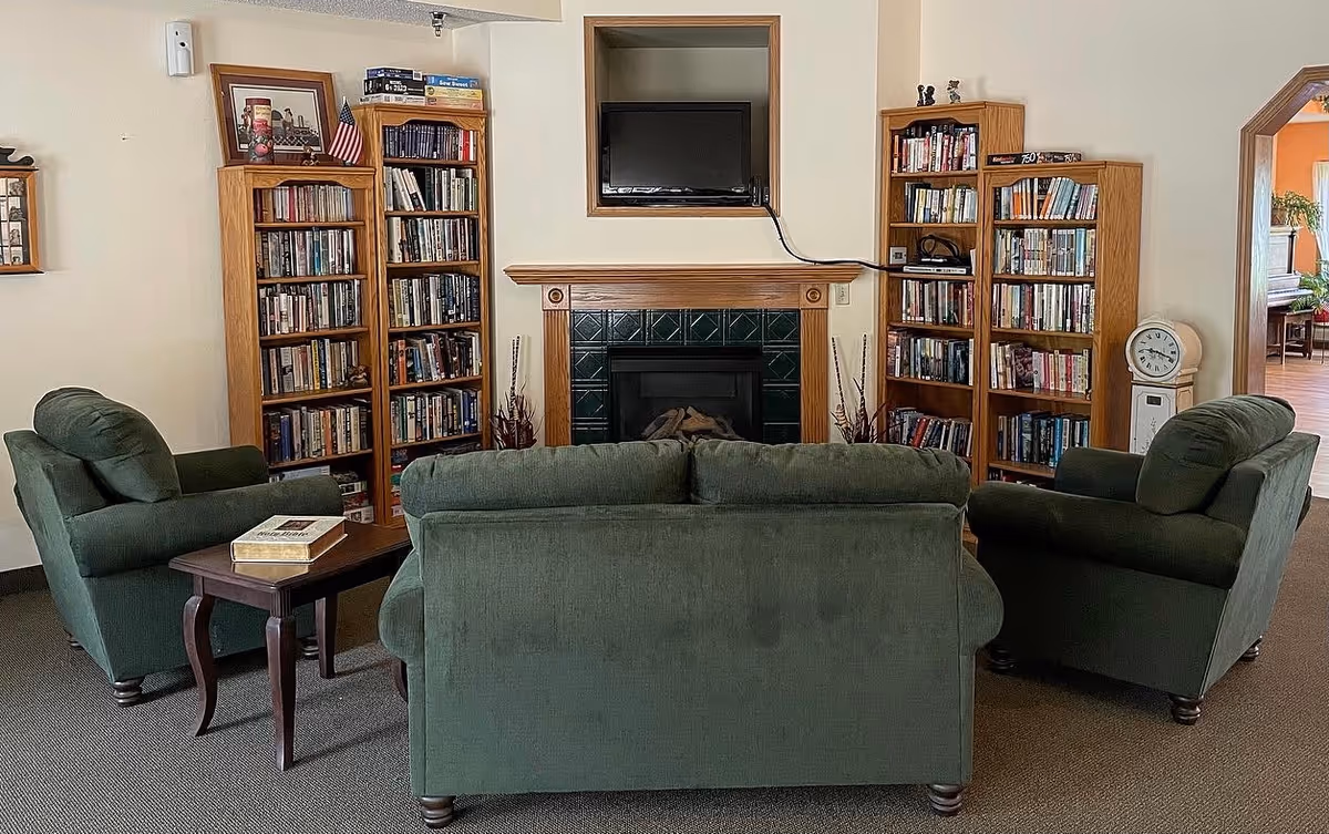 Cozy common room with green upholstered sofa and armchairs arranged around a small table facing a fireplace and wall-mounted TV, flanked by wooden bookshelves.
