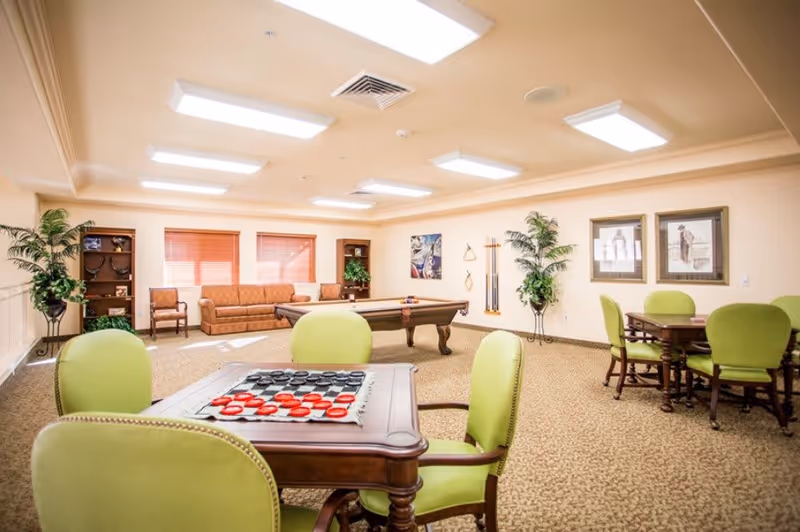 A bright and spacious common room with beige walls and carpet. The room features a pool table in the center, a brown sofa and chairs along the back wall, and two wooden tables with green cushioned chairs. There are plants in decorative stands, framed artwork on the walls, and large ceiling lights providing illumination.