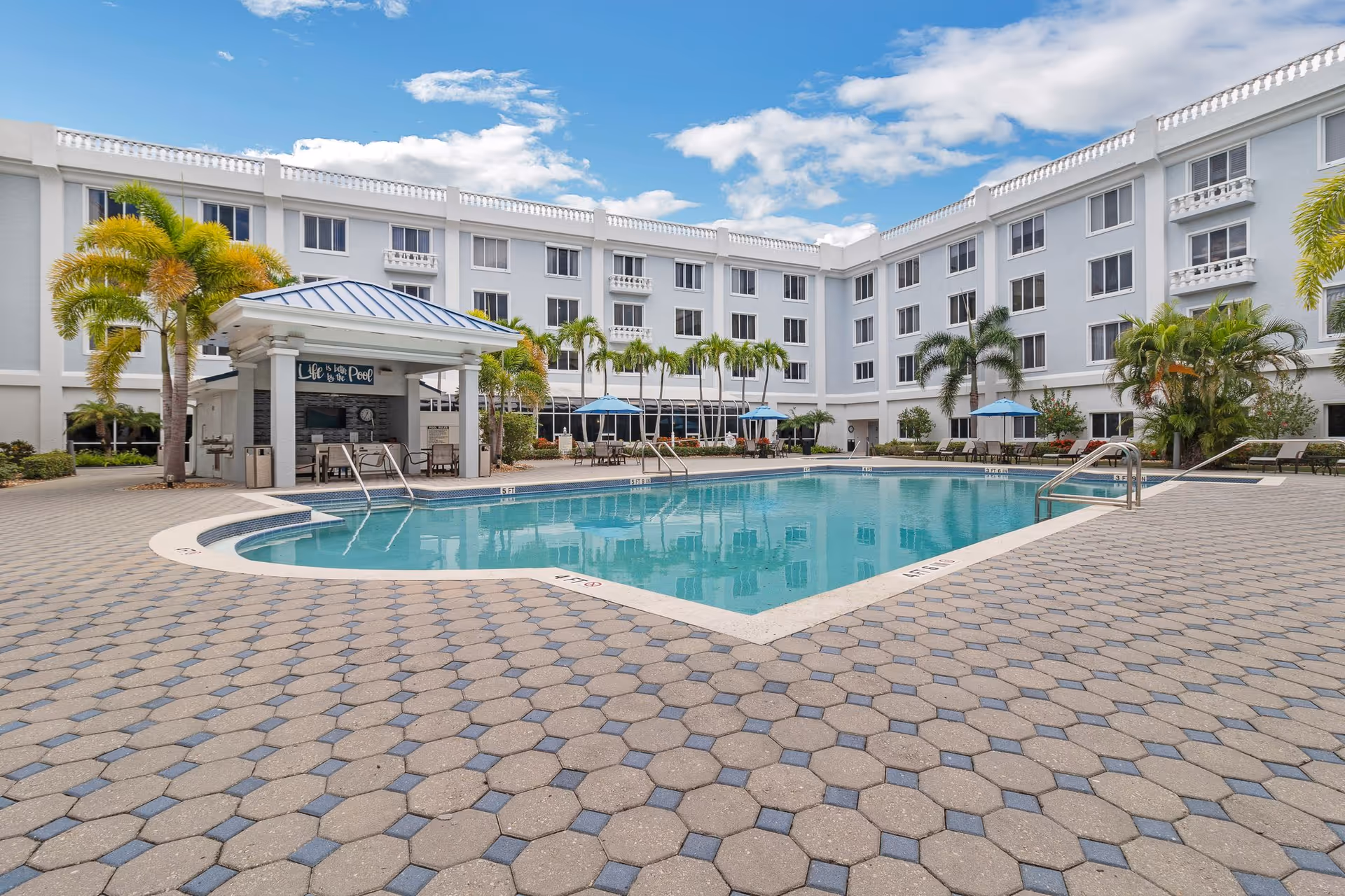 Outdoor swimming pool area at The Summit of Fort Myers with a covered seating area, palm trees, lounge chairs, and umbrellas surrounding the pool. The building with multiple windows and balconies encloses the pool area under a partly cloudy blue sky.