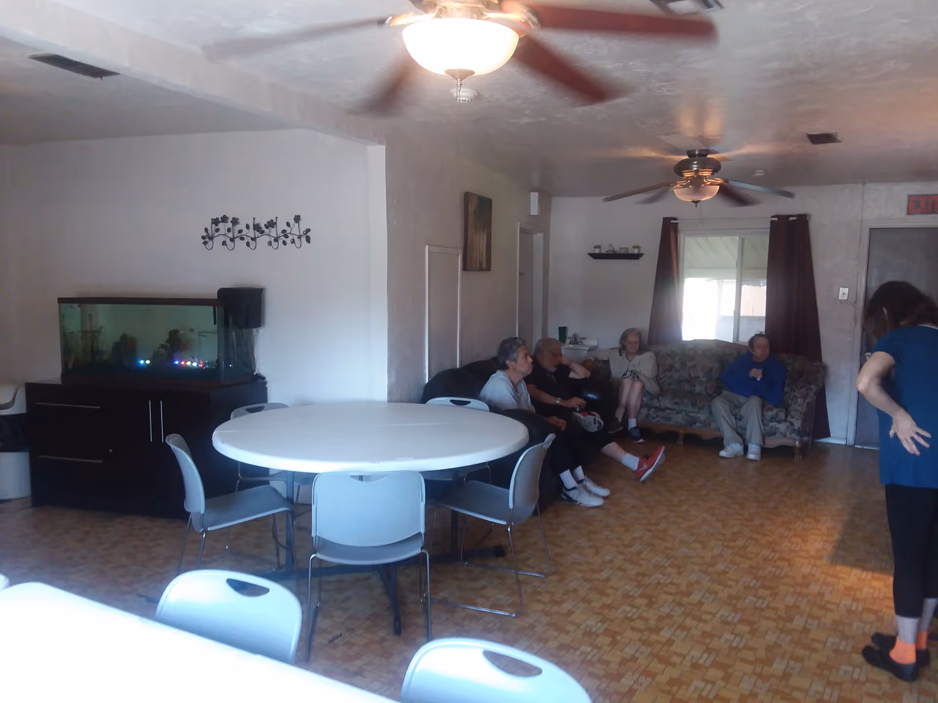A communal living room with round tables and chairs, an aquarium, ceiling fans, and several residents seated on sofas.