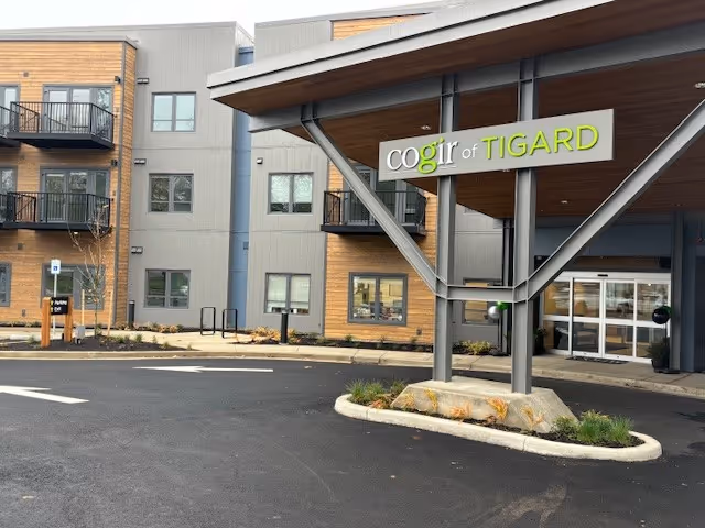 Exterior view of the Cogir of Tigard senior living facility showing the entrance canopy with the facility name sign, a paved driveway, and a multi-story building with balconies and windows.