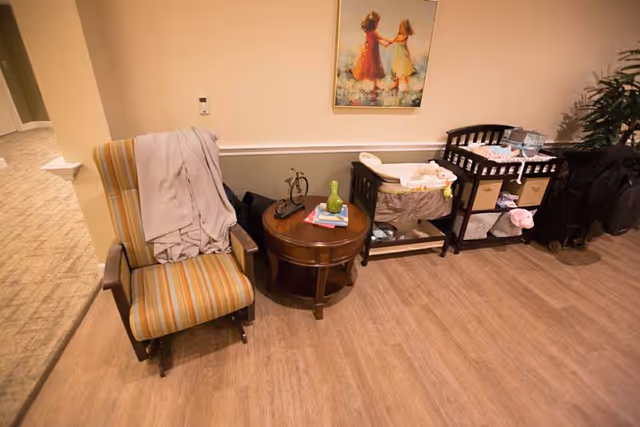 A cozy corner of a room featuring a striped armchair with a light gray blanket draped over it, a round wooden side table with decorative items including a small bicycle sculpture and a green vase, and a dark wooden changing table with baby supplies. A framed painting of two children holding hands hangs on the wall above the changing table. The floor is wood and the walls are painted in two tones with a chair rail.