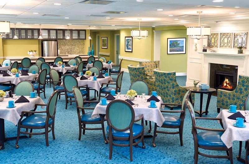 Dining room with multiple round tables set with blue cups and napkins, teal chairs, and a fireplace in the background.