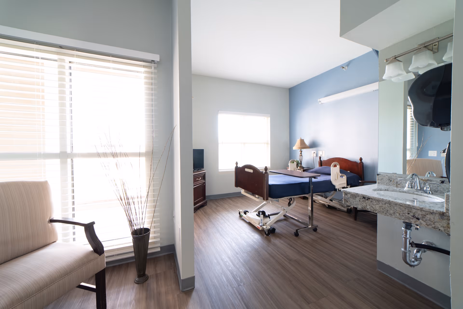 A bright and clean healthcare facility room with a hospital bed, a bedside table with a lamp, a TV on a stand, a chair near a large window with blinds, a tall vase with decorative branches, and a sink with a granite countertop and mirror.