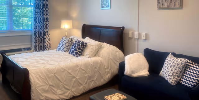 A cozy bedroom featuring a dark wooden bed with white textured bedding and decorative pillows. Next to the bed is a nightstand with a lamp, and a window with patterned curtains allowing natural light to enter. Adjacent to the bed is a dark blue sofa with patterned pillows and a white throw blanket. The walls have framed artwork.
