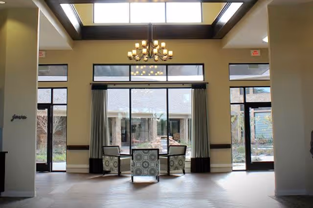 A bright and spacious common area with large floor-to-ceiling windows and glass doors leading outside. Three patterned armchairs are arranged in a semi-circle facing the windows. A modern chandelier hangs from the ceiling, and natural light fills the room.