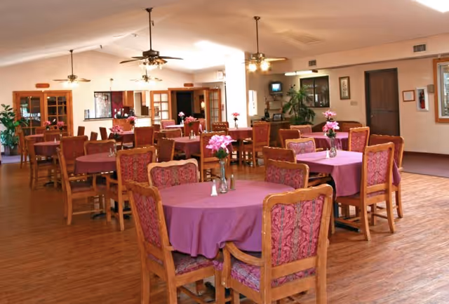 A spacious dining room with multiple wooden tables covered with purple tablecloths and matching wooden chairs with patterned upholstery. Each table has a small vase with pink flowers. The room has wooden flooring, ceiling fans with lights, and large windows allowing natural light. There are plants and framed pictures on the walls, creating a warm and inviting atmosphere.
