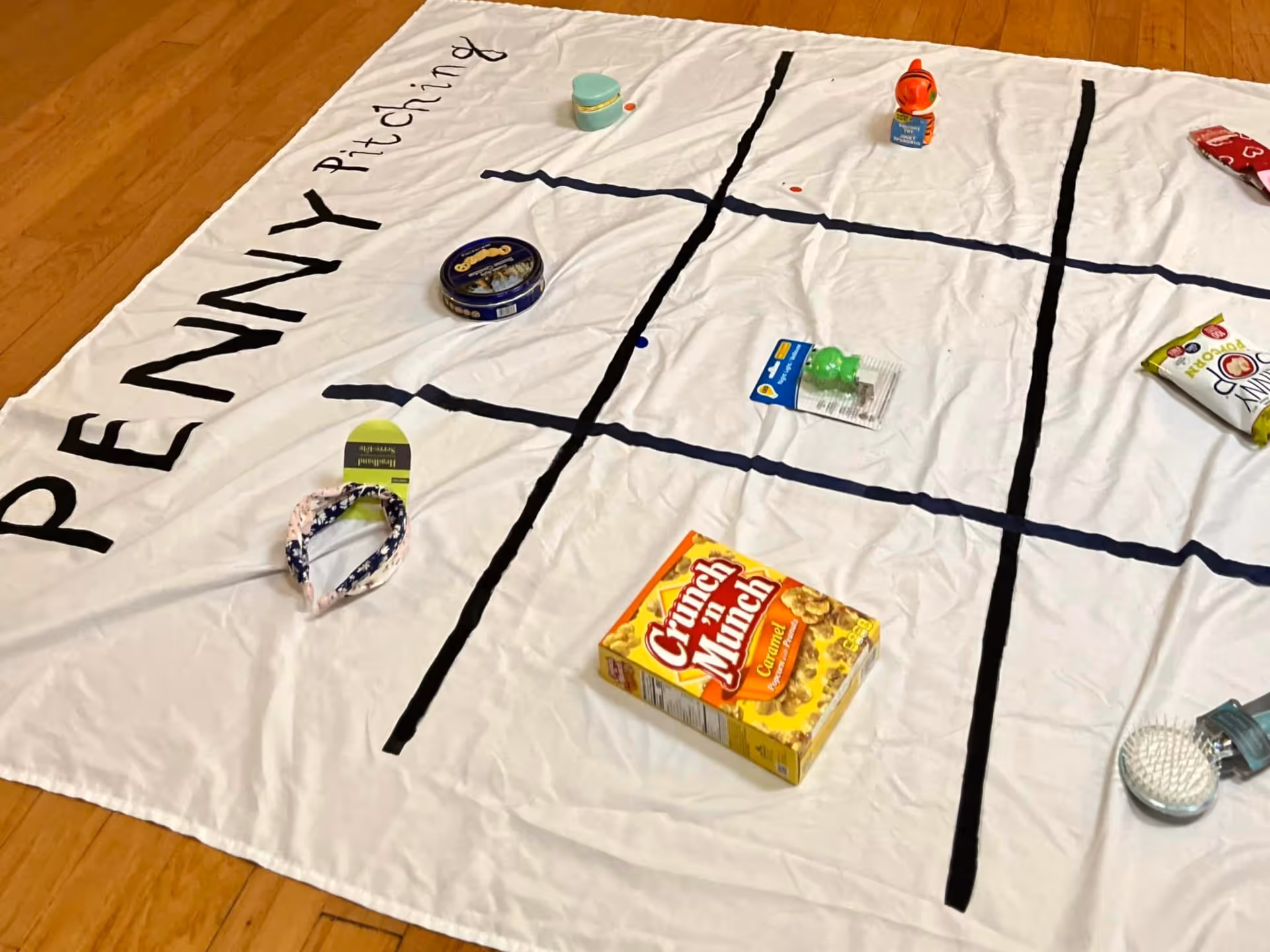 A white cloth laid out on a wooden floor with black lines dividing it into a tic-tac-toe grid. The words 'PENNY Pitching' are written on the cloth. Various small items such as a box of Crunch 'n Munch caramel popcorn, a tin container, a headband, a small toy, and other small objects are placed within the grid squares.