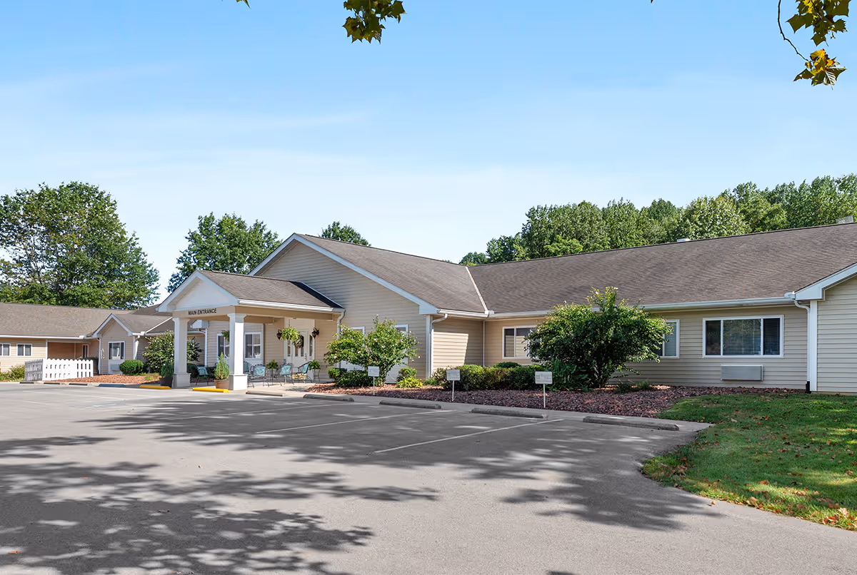 Exterior view of a single-story beige building with a main entrance covered by a small portico supported by white columns. There are several chairs and hanging plants near the entrance, surrounded by landscaped bushes and trees. The parking lot in front is empty, and the sky is clear and blue.