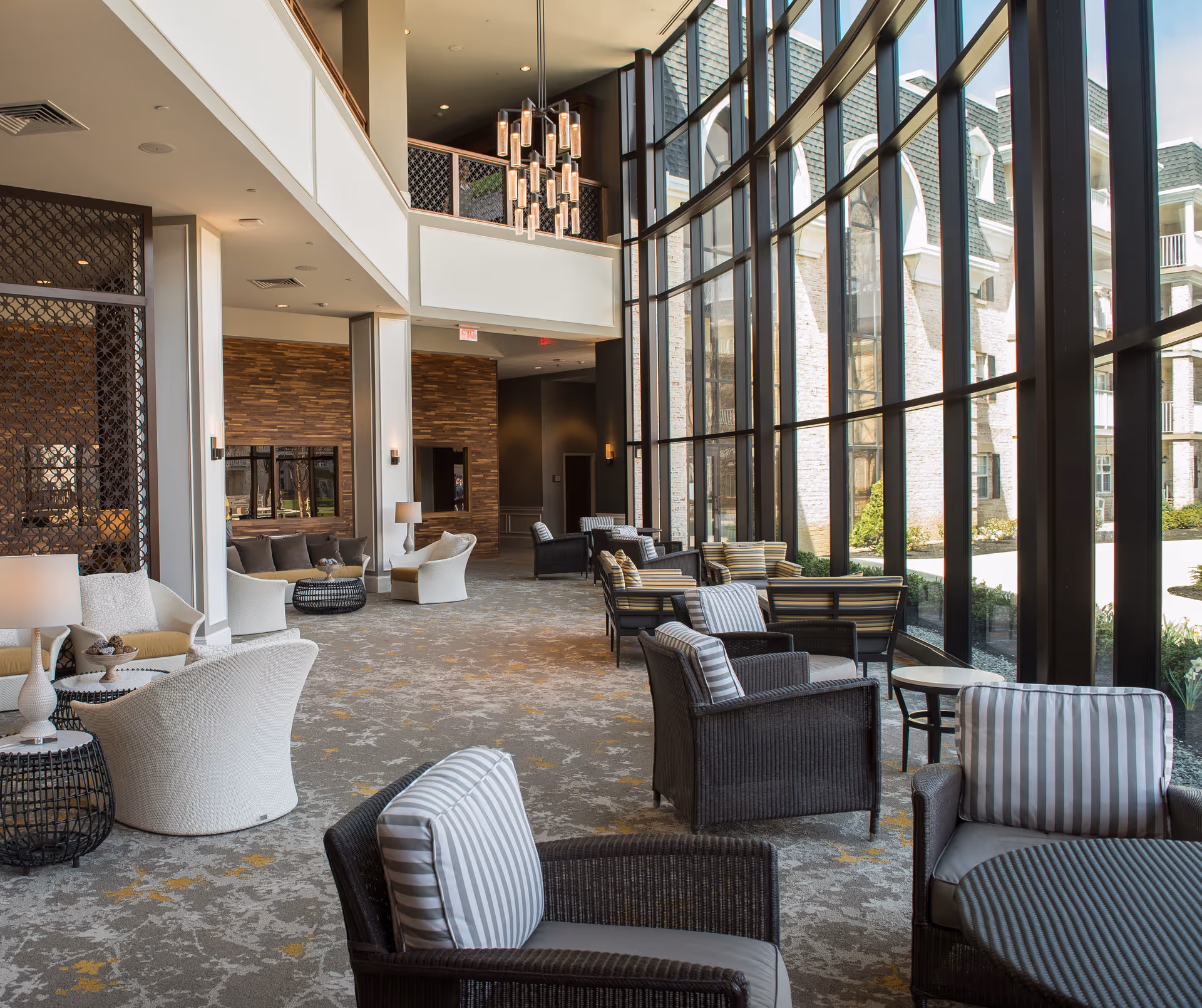 A spacious and well-lit lounge area with large floor-to-ceiling windows allowing natural light to fill the space. The room features multiple seating arrangements with cushioned chairs and small tables, a high ceiling with modern hanging light fixtures, and a carpeted floor with a subtle pattern. The interior design includes neutral tones and wood accents, creating a comfortable and inviting atmosphere.