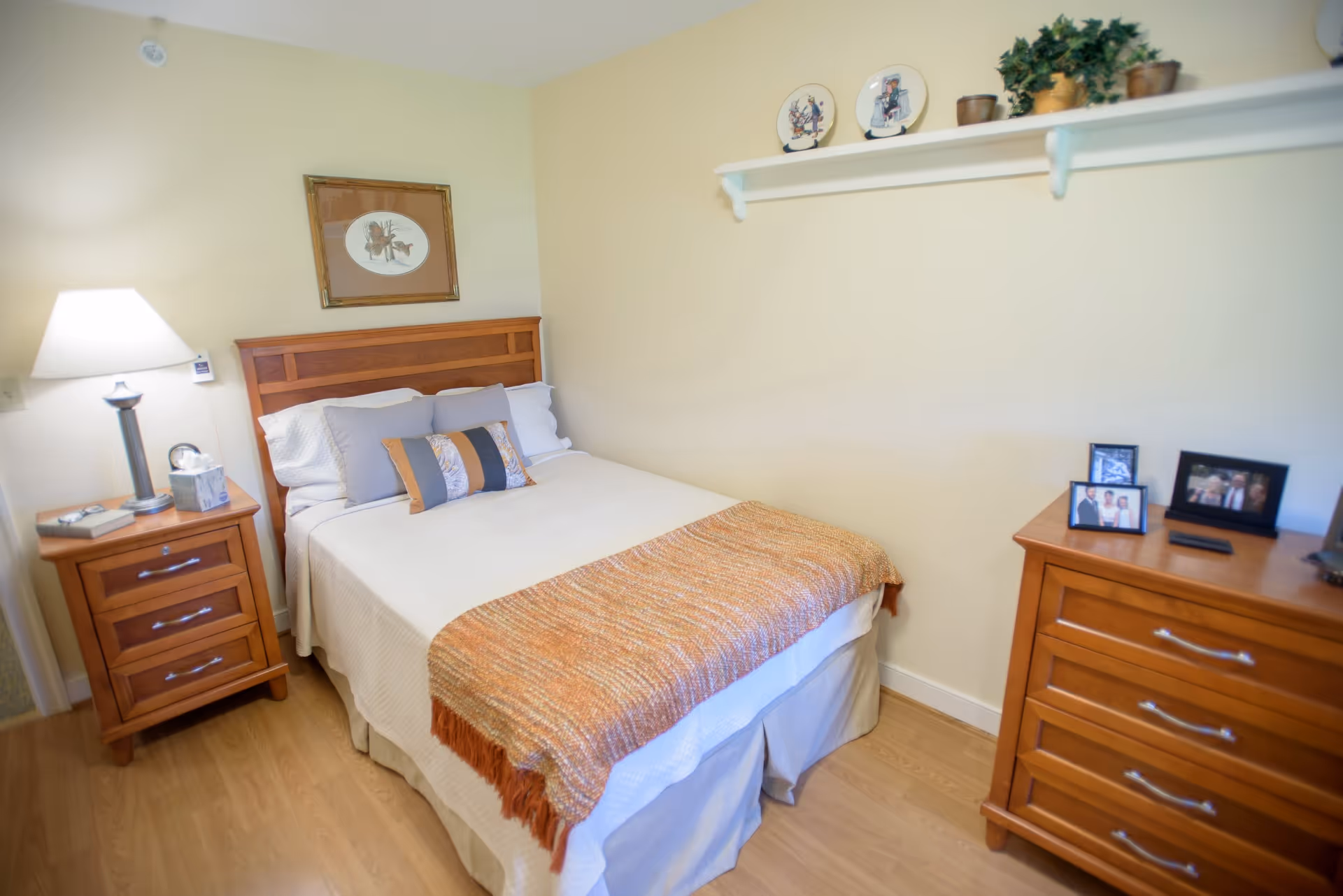 A cozy bedroom with a wooden bed frame and a neatly made bed featuring white and gray pillows and an orange throw blanket. There is a wooden nightstand with a lamp, clock, and tissue box on the left side of the bed. On the right side, there is a wooden dresser with framed photos and a small black device. A white shelf above the dresser holds decorative plates and potted plants. The room has light-colored walls and wooden flooring.