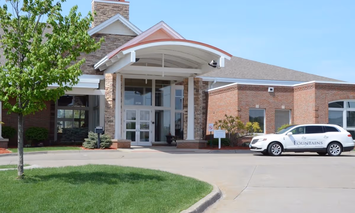 Front exterior view of a brick building with a covered entrance and a white vehicle parked nearby with the word 'Fountains' on its side. There is a tree and green grass in the foreground under a clear blue sky.