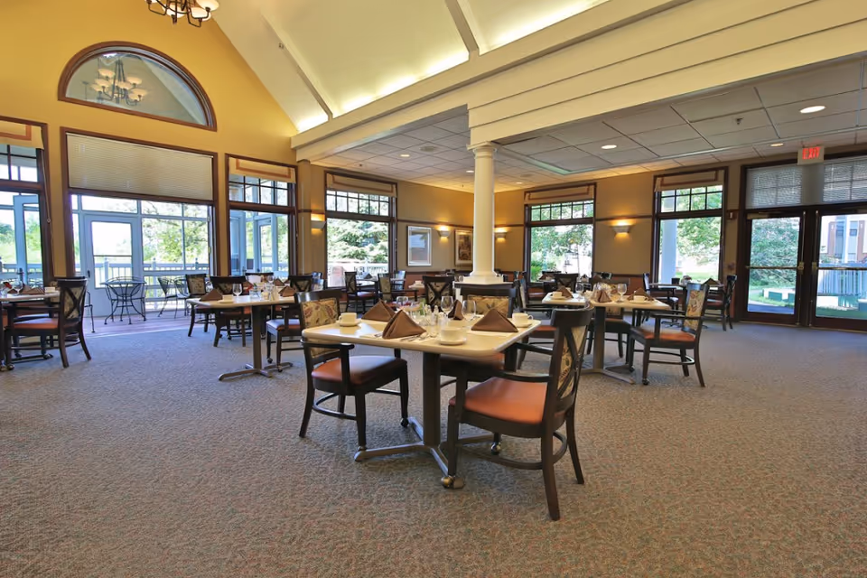 A spacious dining room in a senior living facility with multiple tables set for meals, each with folded napkins, cups, and glasses. The room features large windows allowing natural light to fill the space, a high vaulted ceiling with a chandelier, and a carpeted floor. There is an outdoor patio visible through the windows.
