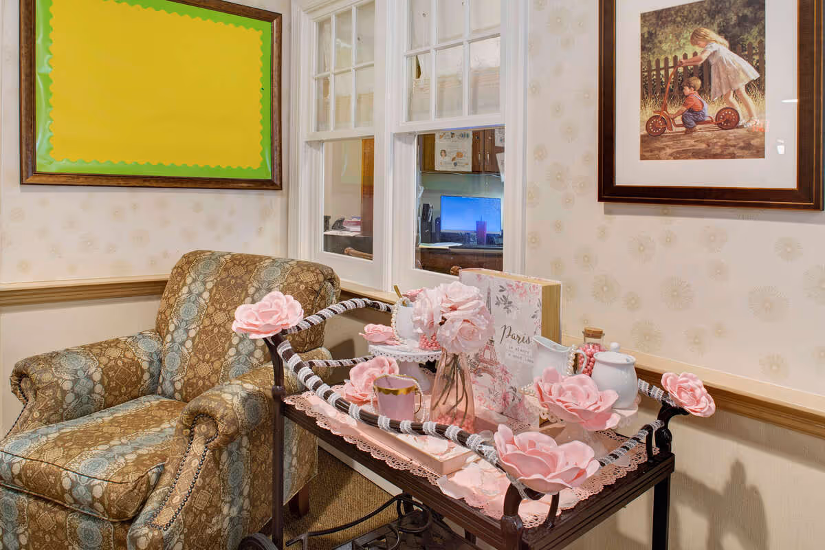 A cozy corner in a senior living facility featuring a patterned armchair next to a decorative cart adorned with pink artificial flowers, a teapot, cups, and a book titled 'Paris'. The wall behind has a framed picture of two children playing and a blank bulletin board with a green border. A window reveals a glimpse into an office area with a computer monitor.