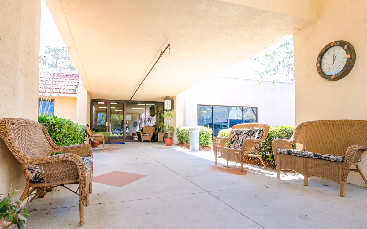 Covered entrance walkway of a nursing facility with wicker chairs and benches under a canopy leading to glass doors.