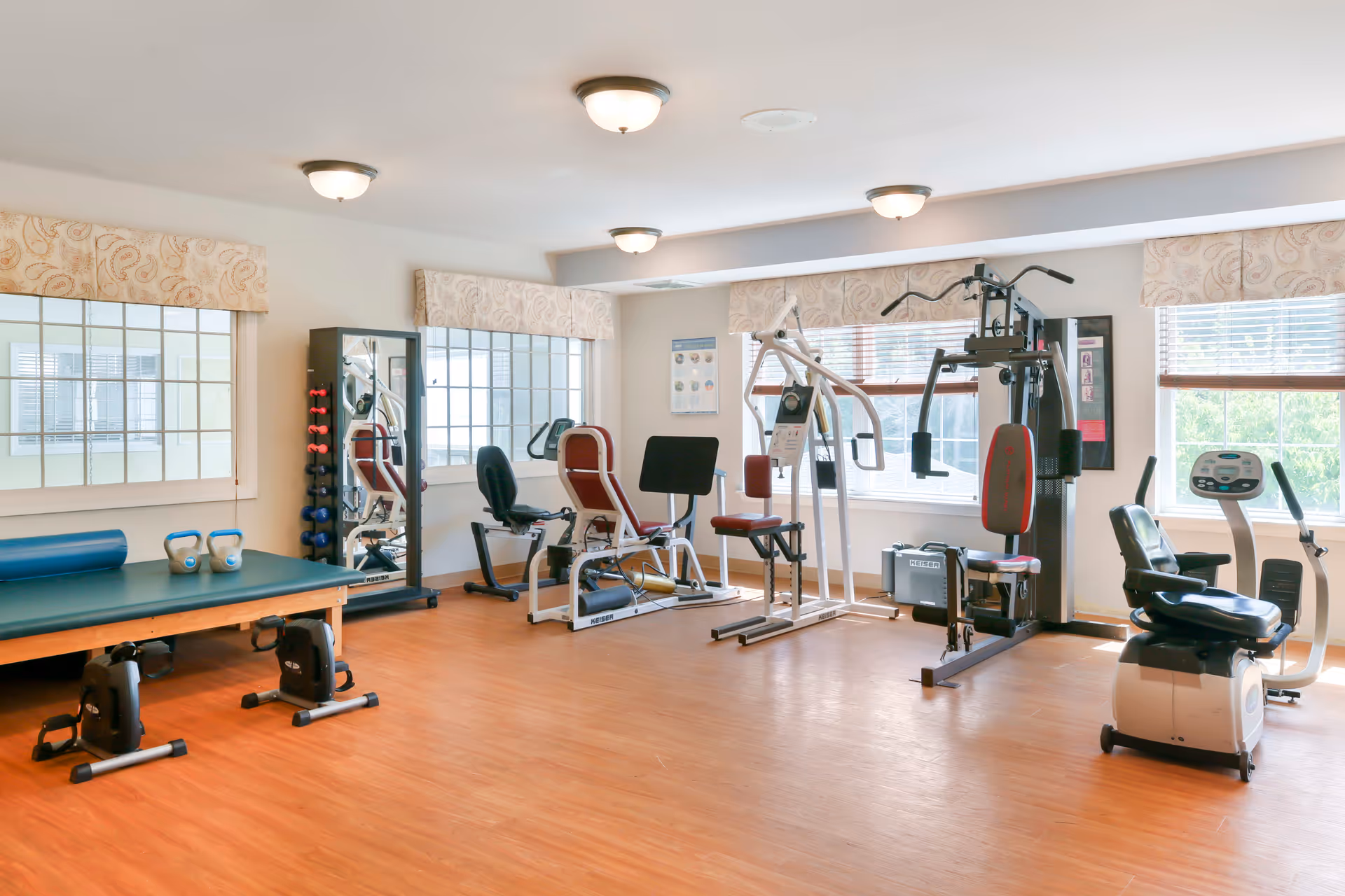 A bright and spacious exercise room with wooden flooring, featuring various fitness equipment including stationary bikes, weight machines, dumbbells, kettlebells, and a padded exercise table. Large windows with patterned valances allow natural light to fill the room.