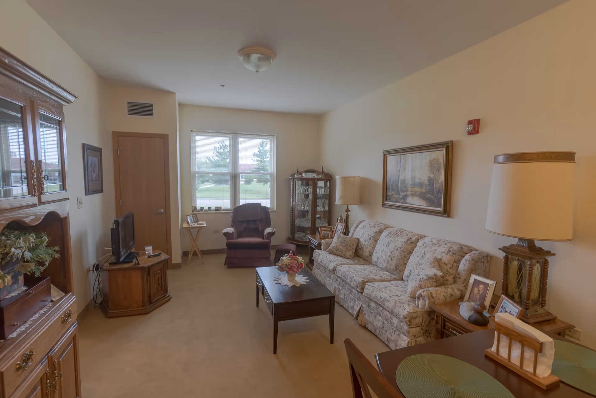 A cozy living room in a senior living facility with a floral-patterned sofa, a wooden coffee table with a flower vase, a burgundy recliner chair near a window, a wooden cabinet with glassware, a TV on a wooden stand, framed pictures on side tables, and a large lamp. The room has beige walls and carpeted floor.