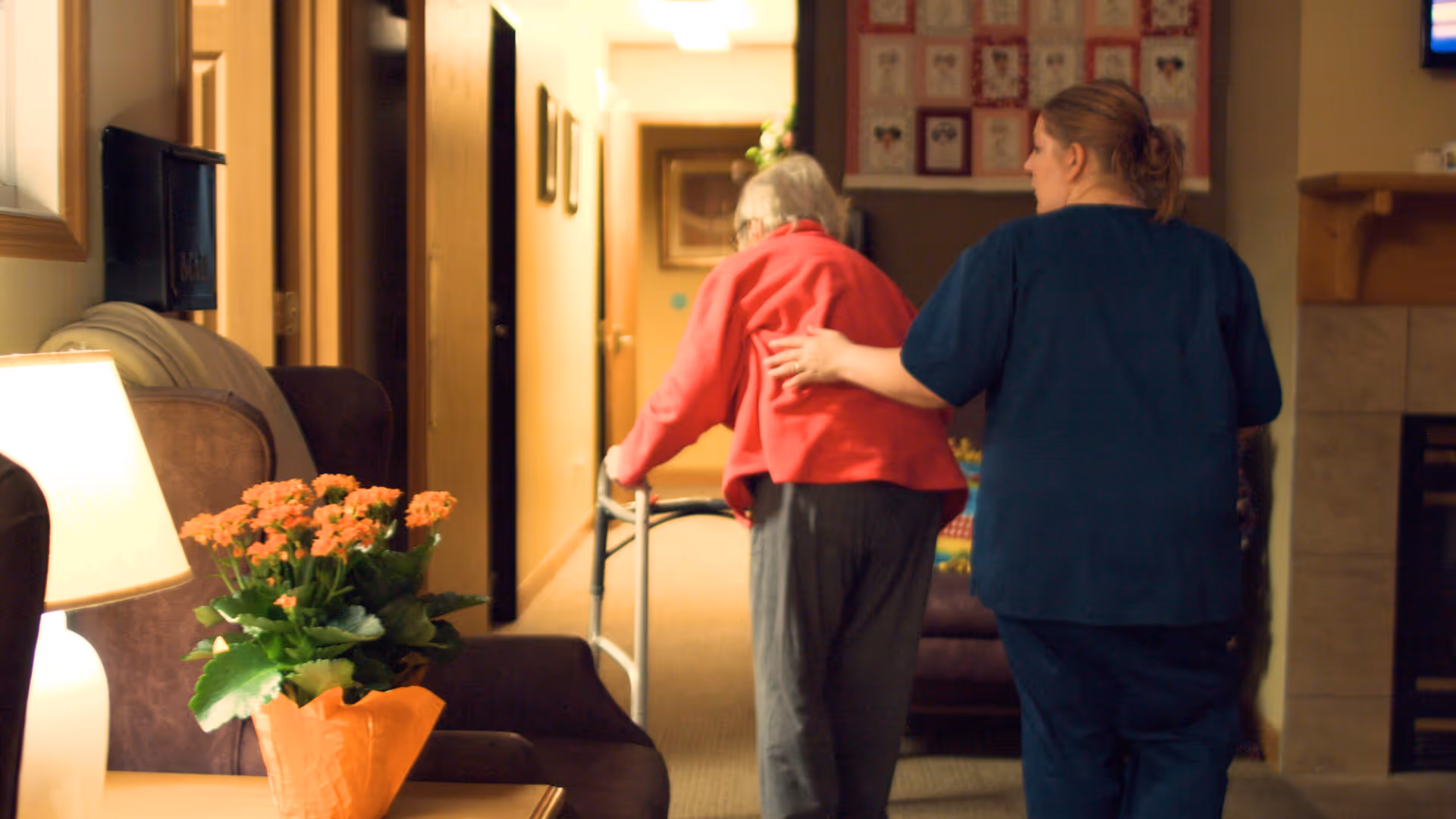 A caregiver in blue scrubs assists an elderly woman using a walker as they walk down a hallway in a cozy living area with a lamp and a potted plant on a table nearby.