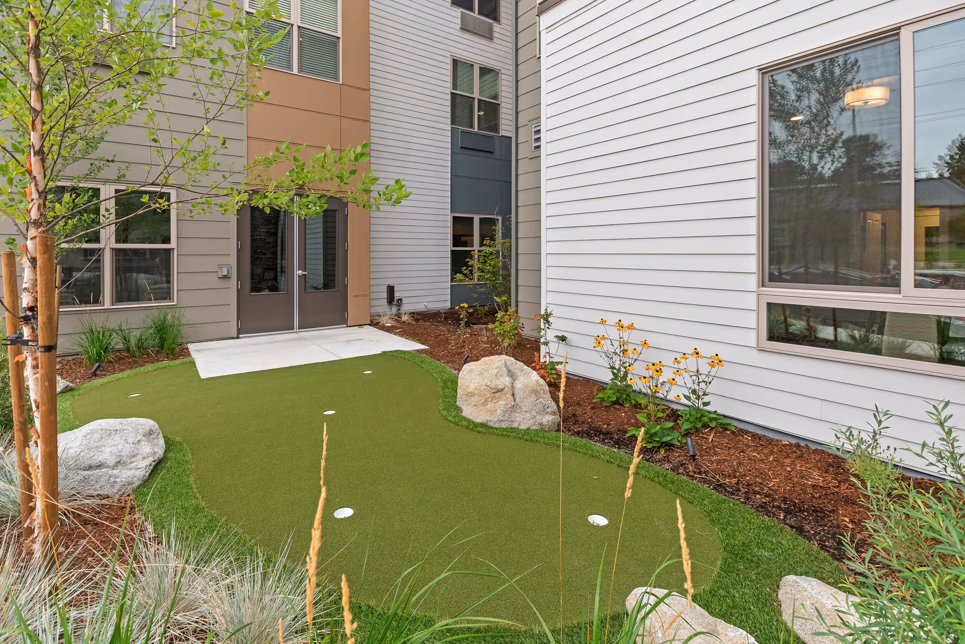 Outdoor courtyard area with a small artificial putting green surrounded by landscaping including rocks, plants, and trees. The area is adjacent to a building with large windows and double doors.