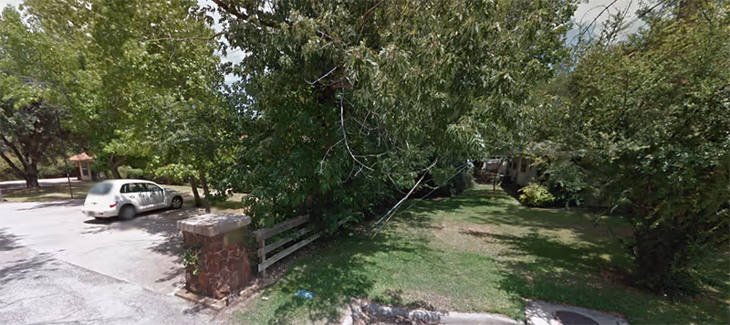 Tree-lined driveway and grassy front yard with a stone pillar near a parked car.