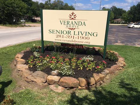A large outdoor sign for Veranda House Senior Living surrounded by a circular flower bed with rocks and various plants, located next to a road with cars and trees in the background.