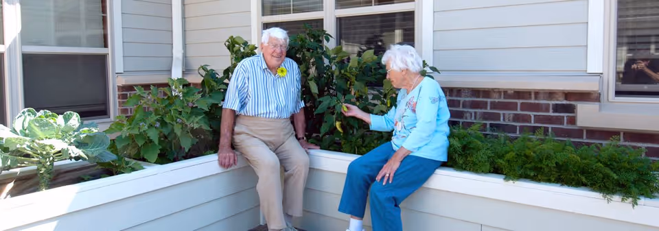 Two elderly residents sitting on the edge of a raised planter in a sunny courtyard beside shrubs and a building.