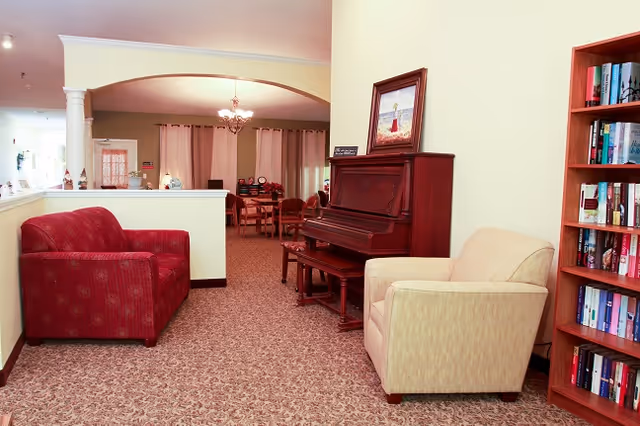 Interior view of a senior living facility lounge area with a red patterned loveseat on the left, a beige armchair on the right, a wooden upright piano with a framed painting on top, and a bookshelf filled with books. In the background, there is a dining area with tables and chairs under a chandelier, and windows with curtains.