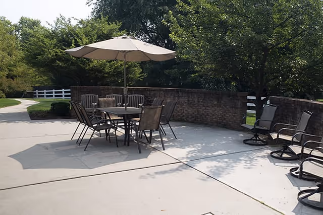 Outdoor patio area with a table, six chairs, and a large umbrella providing shade. Additional chairs are arranged along a curved brick wall, surrounded by trees and greenery.