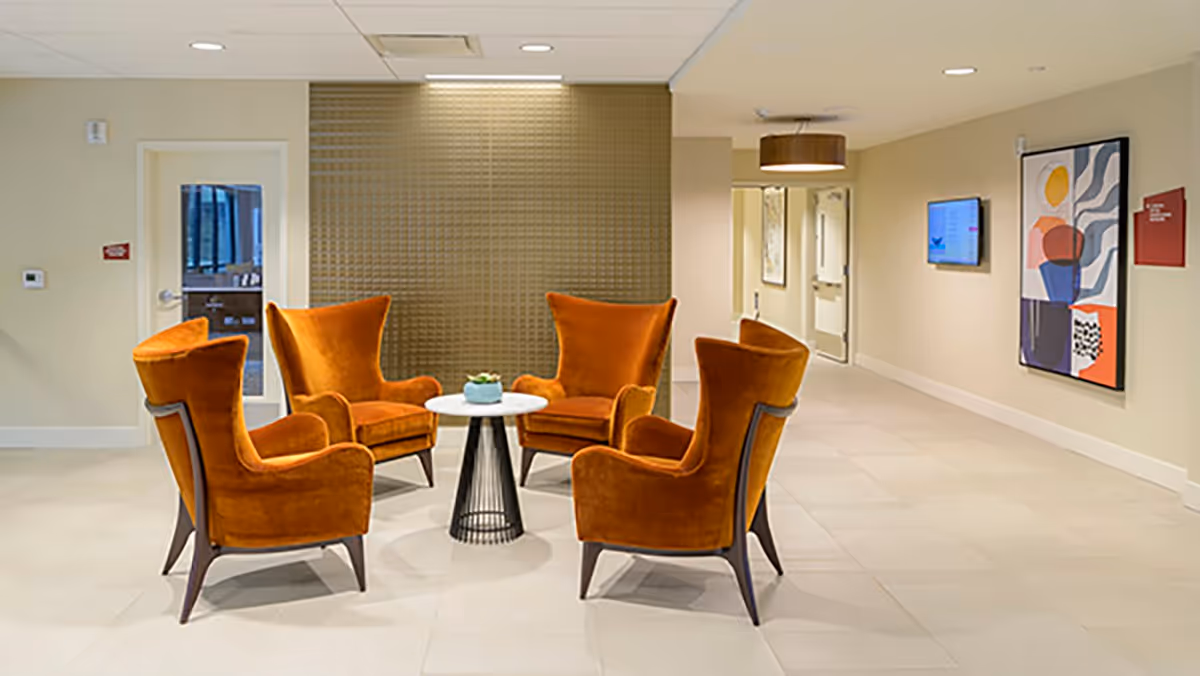 A seating area in a senior living facility with four orange upholstered armchairs arranged around a small round white table with a small plant on it. The room has beige walls, a textured accent wall, a hallway with doors, a wall-mounted TV, and modern artwork on the wall.