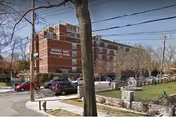 Front exterior of Morris Park Rehabilitation & Nursing Center, a multi-story brick building with parked cars, a lawn and stone fence.