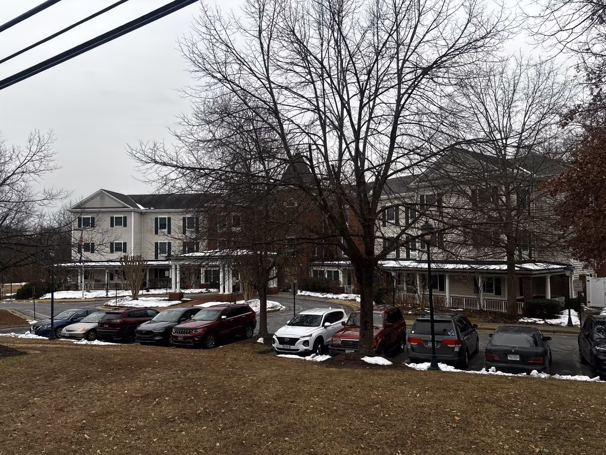 Front exterior of a multi-story senior living building with parked cars, leafless trees, and patches of snow on the ground.