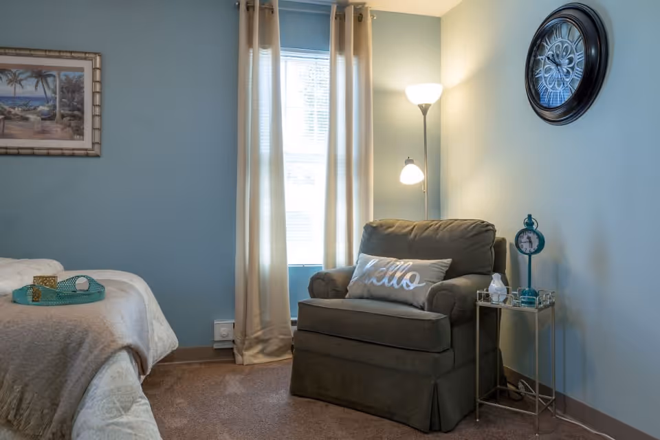 A cozy corner of a bedroom featuring a comfortable armchair with a 'Hello' pillow, a small side table with a decorative clock and a lamp, a floor lamp providing warm light, a window with beige curtains, and part of a bed with a tray on it. The walls are painted light blue and there is a framed beach-themed picture hanging on the wall.