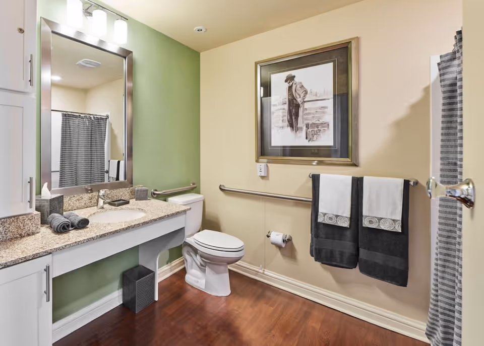 Well-lit accessible bathroom with a granite-topped vanity and mirror, toilet, grab bars, towel rack, and a shower with a striped curtain.