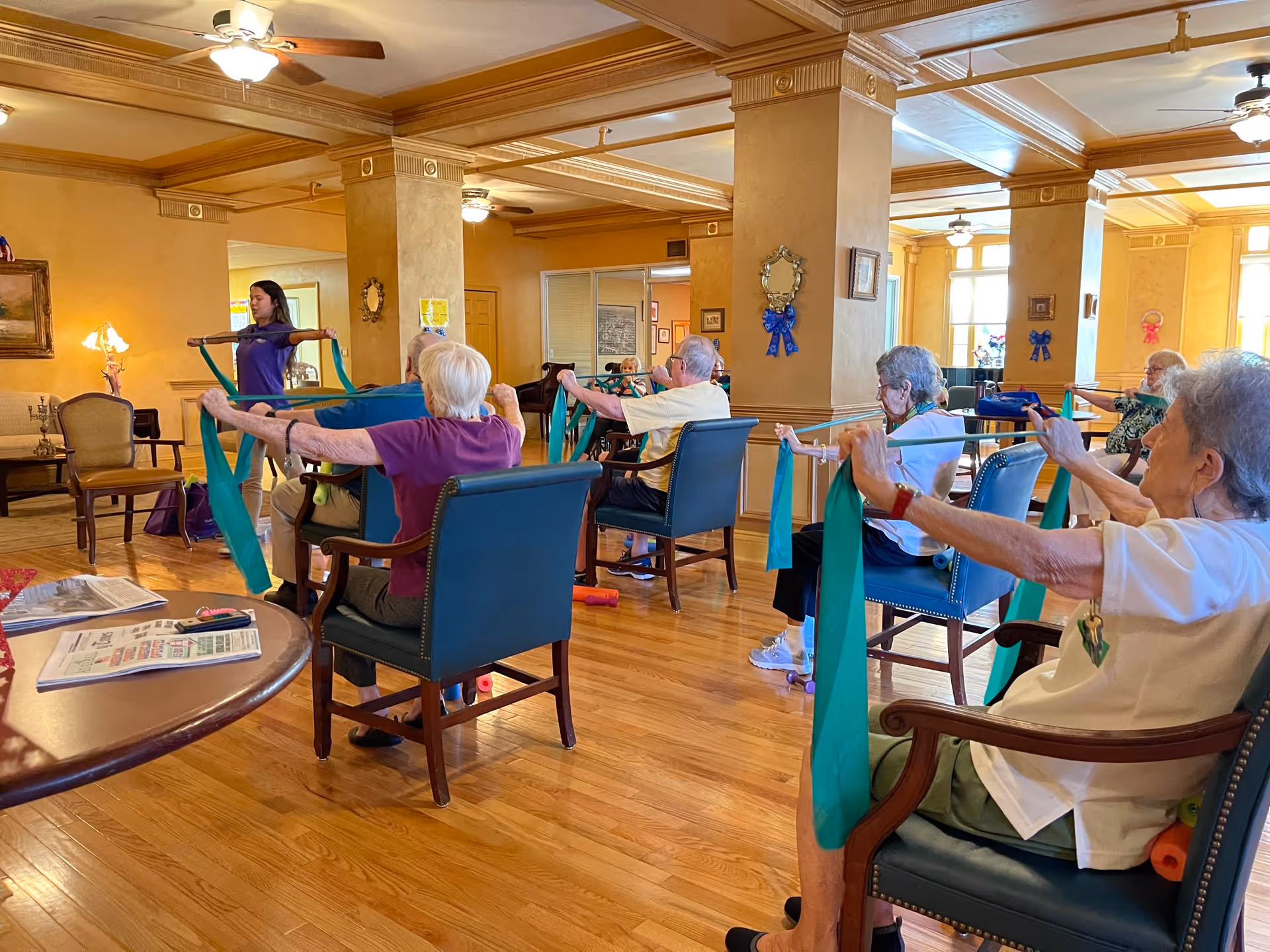 A group of elderly individuals seated in a spacious, well-lit room participating in a seated exercise class using green resistance bands, led by a young instructor standing at the front. The room has wooden floors, ceiling fans, and warm yellow walls with decorative molding and framed pictures.