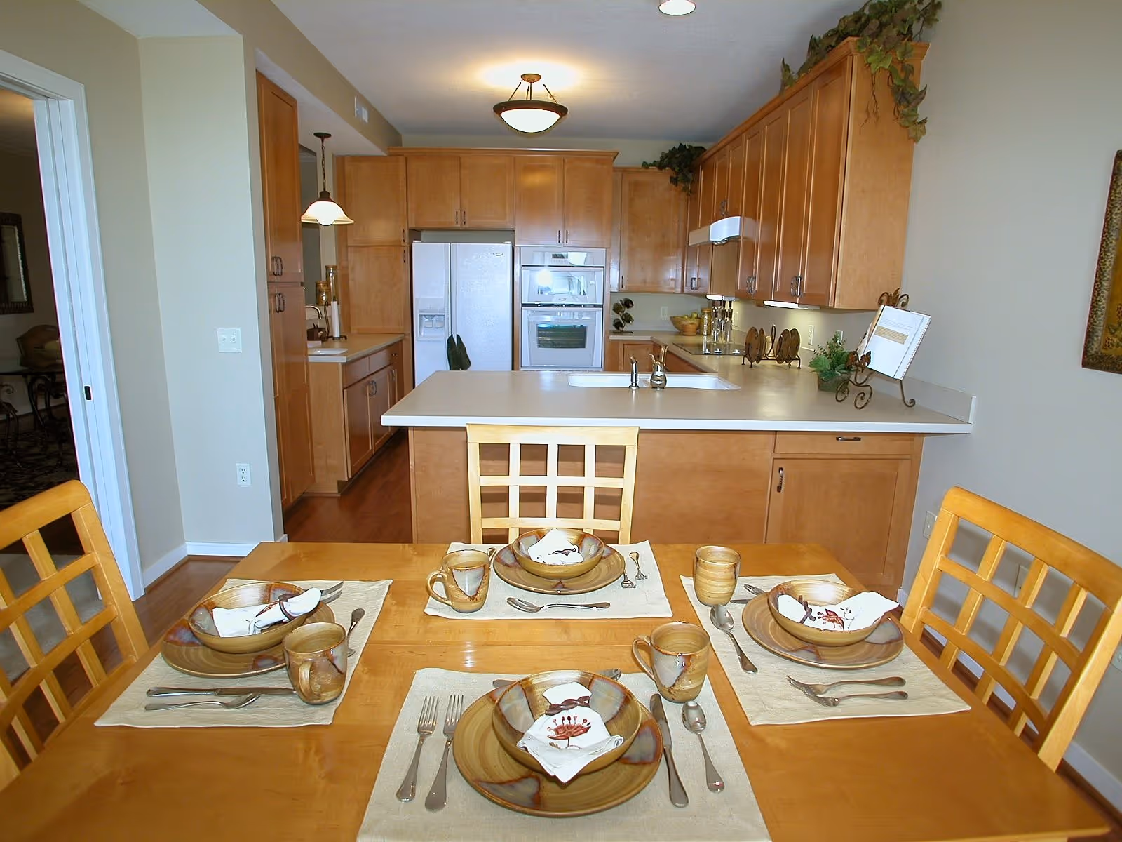 A dining table set for four with plates, cups, and silverware in the foreground, and a kitchen with wooden cabinets, a refrigerator, oven, and a sink in the background.