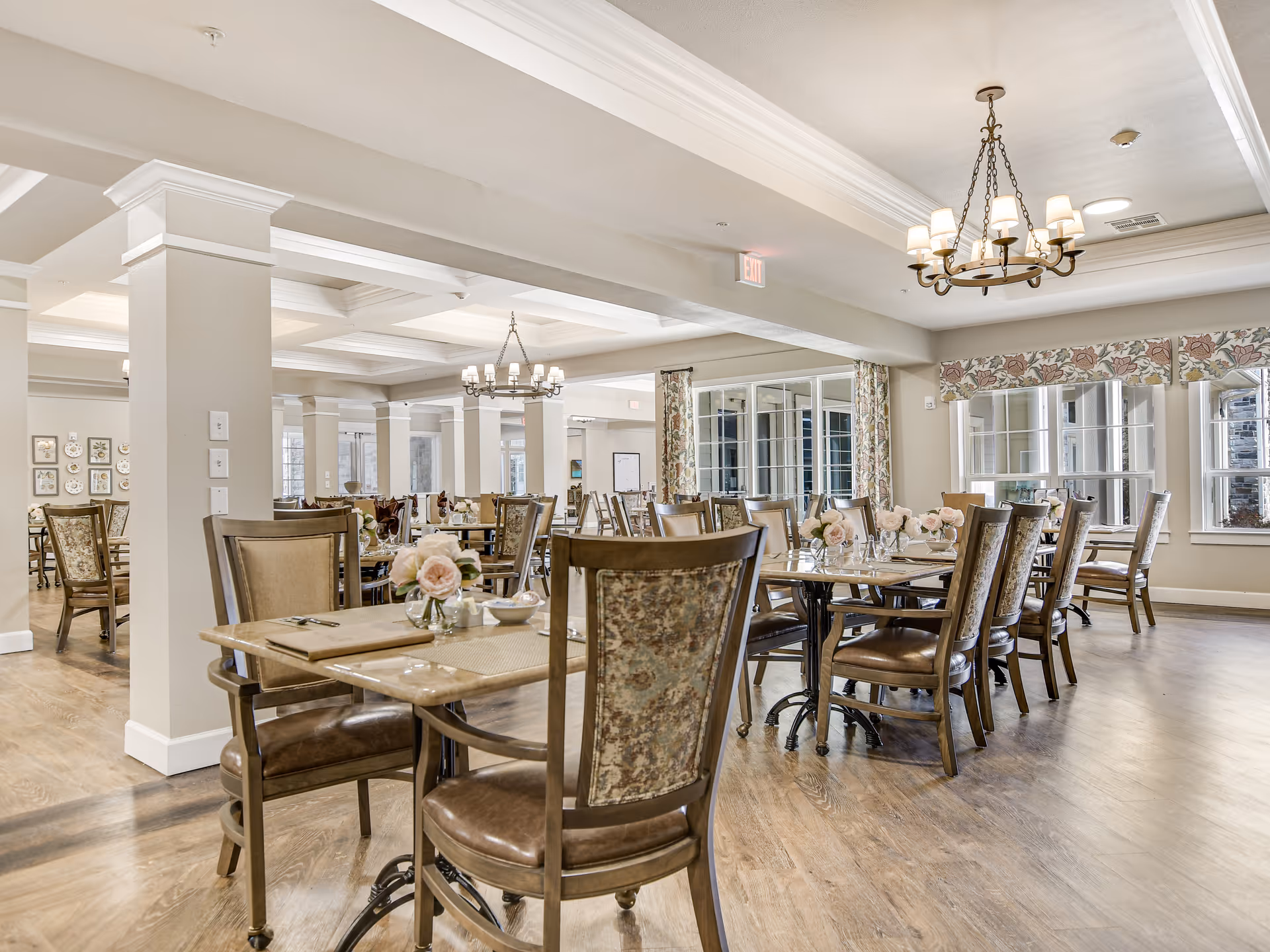 A spacious and well-lit dining room with multiple wooden tables and chairs arranged neatly. Each table is decorated with small vases of pink flowers and place settings. The room features large windows with floral curtains, chandeliers hanging from the ceiling, and light-colored walls and ceiling with crown molding. The floor is wooden, and the overall ambiance is warm and inviting.