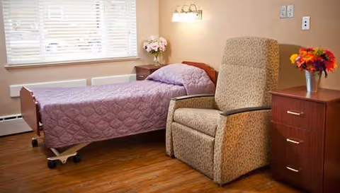 A bedroom in a senior living facility featuring a purple quilted bed, recliner, and dresser.